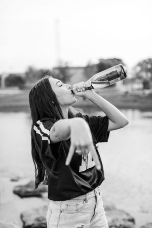 A person in a sports jersey drinks from a glass bottle outdoors in a black and white photograph.