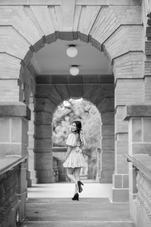 Black and white photo of a figure in a light dress twirling under a stone archway with elegant architectural details.