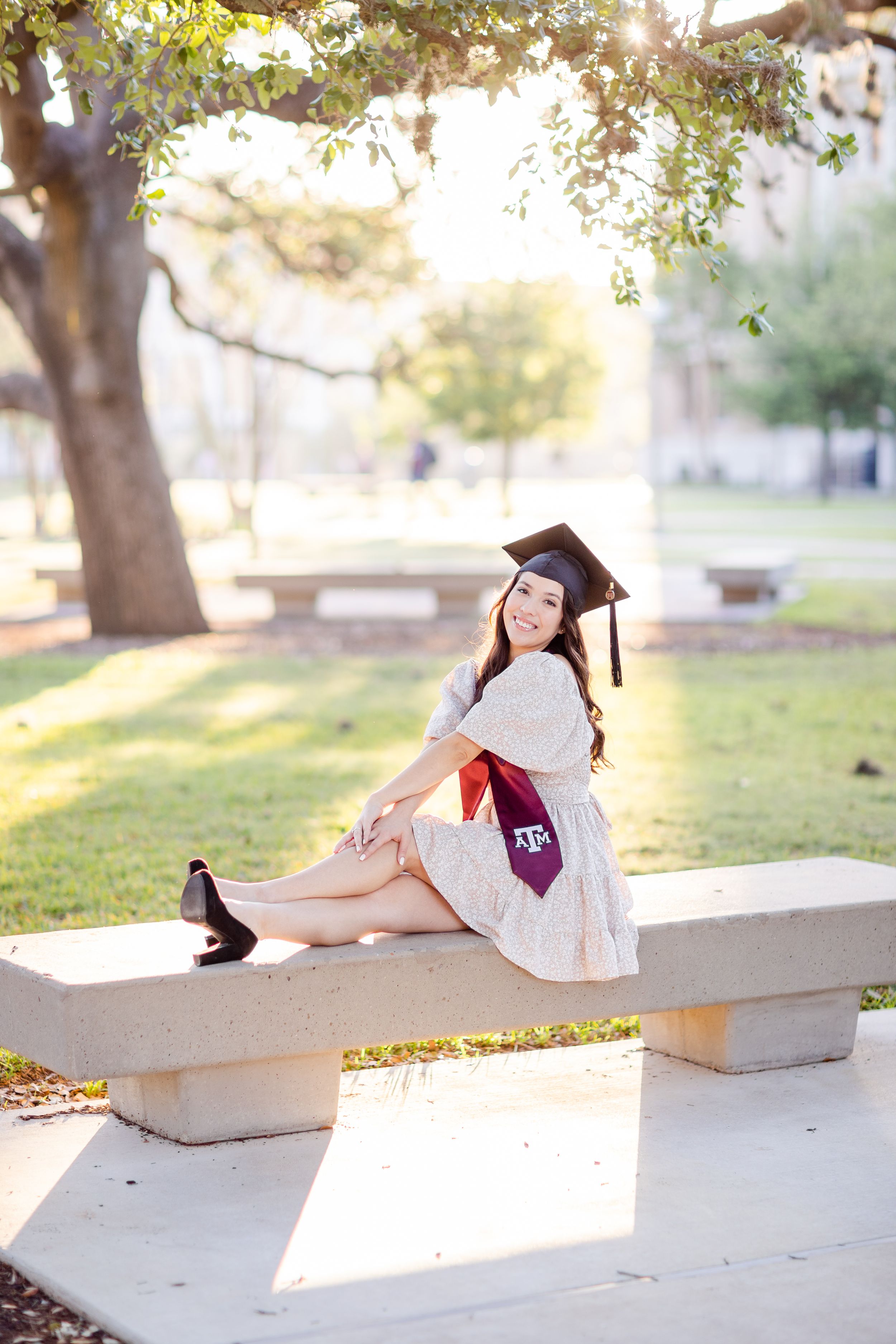 Graduate wearing cap and gown poses on concrete bench in sunny park at Texas A&M campus.