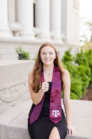 Graduate in a black dress and maroon graduation sash poses by white columns for graduation photos at Texas A&M.