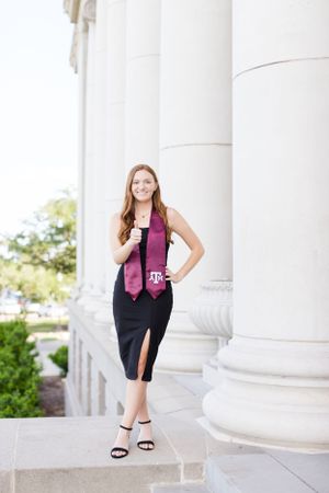A graduate poses between classic white columns wearing a maroon Texas A&M sash and black dress.