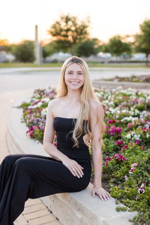 Person in black strapless dress sits on stone ledge near colorful flower garden at sunset.