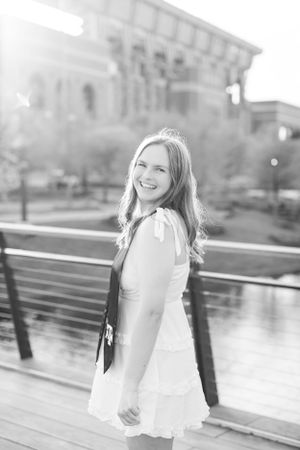 A black and white portrait of someone in a white dress smiling on a wooden deck with a building in the background.