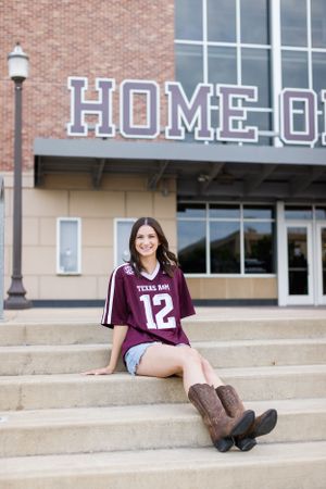 College student in Texas A&M jersey sitting on concrete steps wearing brown boots near a 'HOME OF' sign.