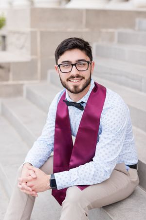 Smartly dressed figure in a burgundy vest, bow tie and glasses sits casually on concrete steps outdoors.