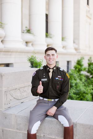 A military cadet in dress uniform gives a thumbs up while sitting on stone steps outside a building with columns.