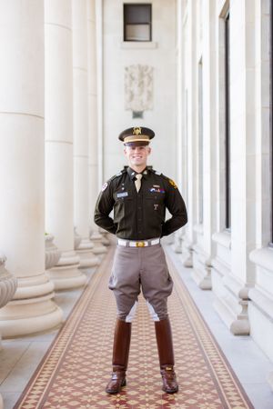 Military officer in dress uniform stands in ornate hallway with white columns and patterned tile floor.