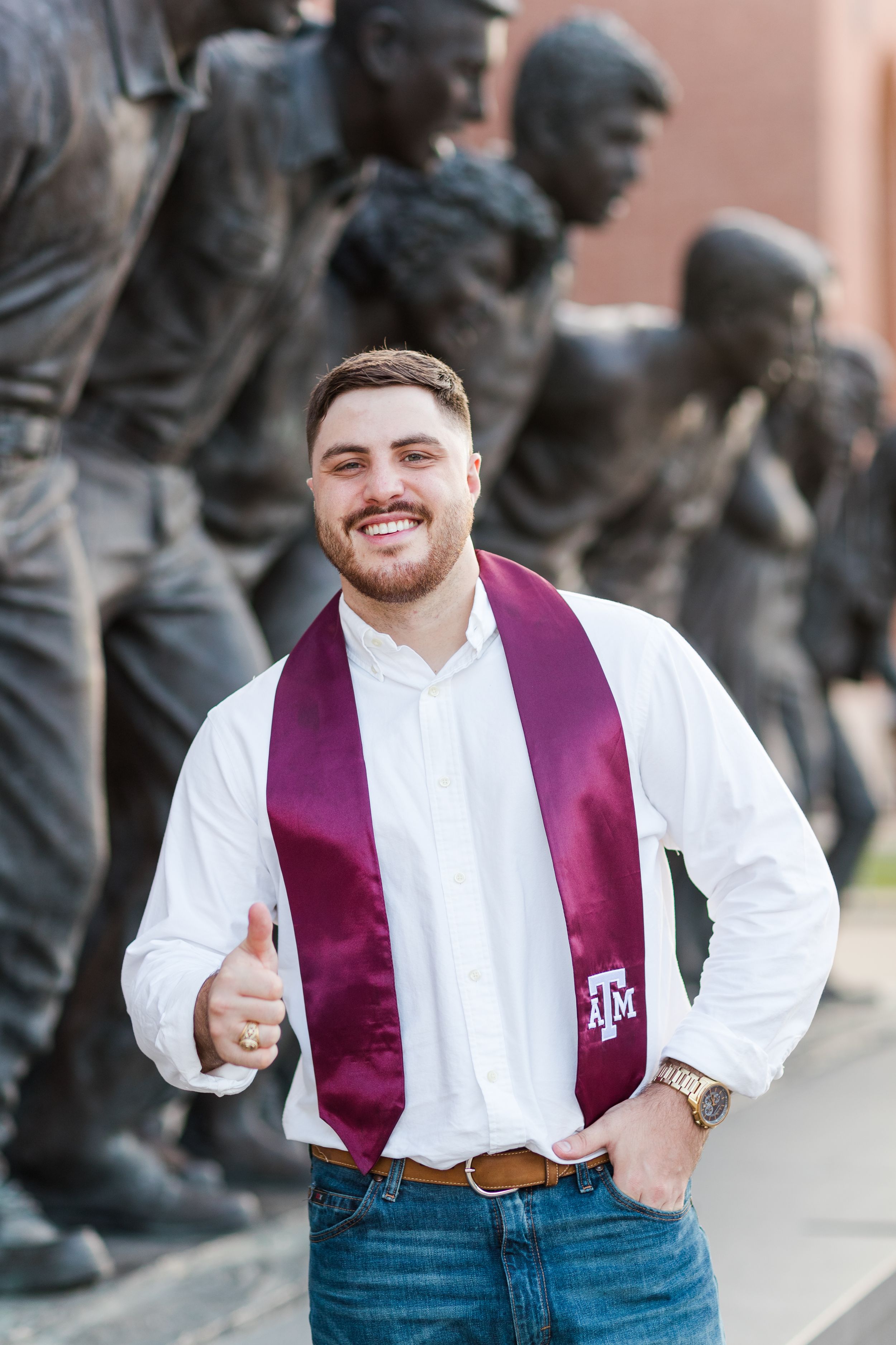 Graduate wearing maroon Texas A&M stole and white shirt gives thumbs up in front of campus statue monument.