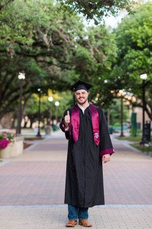 Graduate in black cap and gown with pink stole stands on brick pathway under trees during outdoor graduation photoshoot.