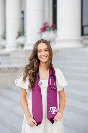 A medical professional wearing a white dress and purple Texas A&M graduation stole with stethoscope stands outside a building.