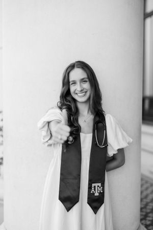 Texas A&M graduate in white dress and graduation stole with stethoscope stands smiling by white wall in black and white photo.
