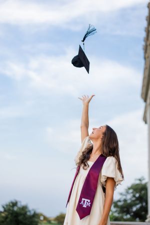 College graduate wearing Texas A&M stole throws graduation cap into blue sky during celebratory photo.