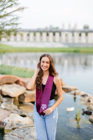 A person wearing a purple tank top and jeans stands by a lake with rocks and a bridge in the background.