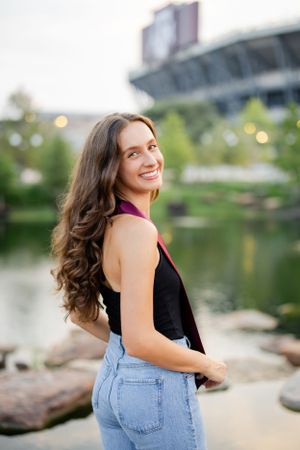 A high school senior wearing a black sleeveless top and jeans poses near a pond at dusk with a stadium in the background.