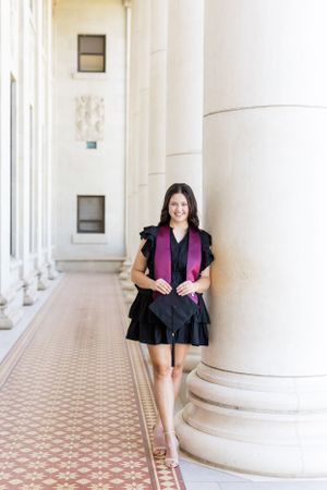 Figure in a black dress and magenta vest stands elegantly in a colonnade with ornate tiled flooring and white columns.