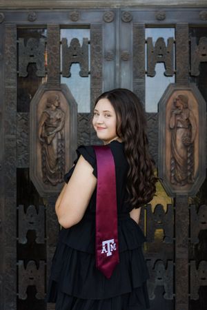 Graduate in black dress poses with Texas A&M maroon tie in front of ornate architectural details with carved statues.