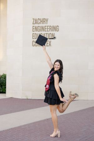 Graduate celebrates outside the Zachry Engineering Education Complex in a black dress, holding up her graduation cap.