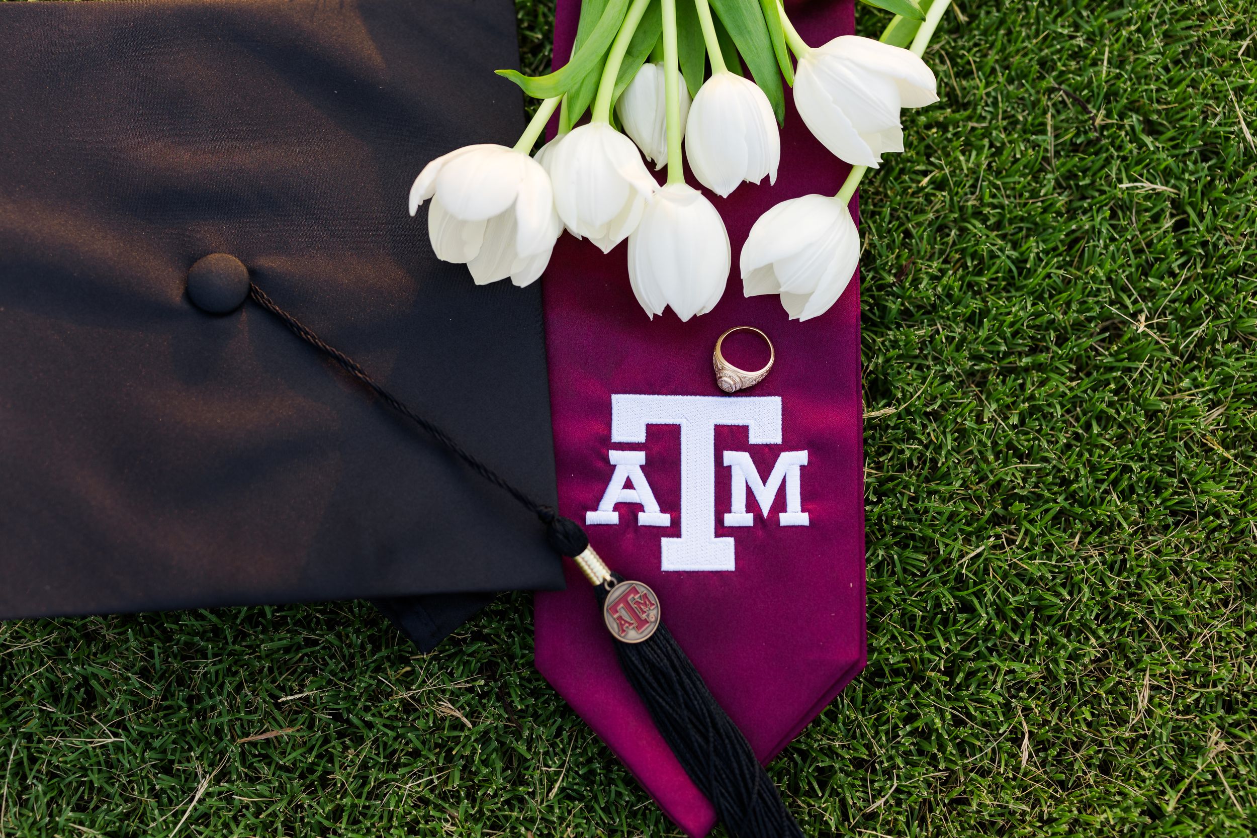 A Texas A&M graduation cap with white tulips and maroon decor on artificial grass.
