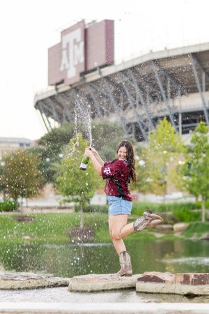 Joyful figure in red plaid shirt and jean shorts splashes water at Texas A&M University campus on a sunny day.