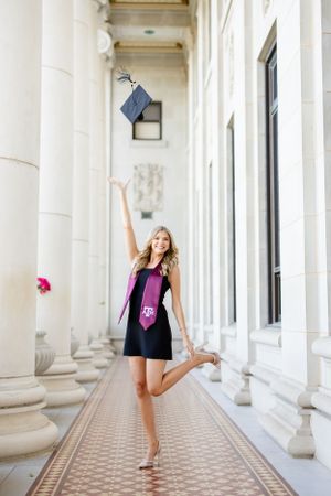 A graduate celebrates with cap toss in a classical columned hallway wearing purple and black dress.