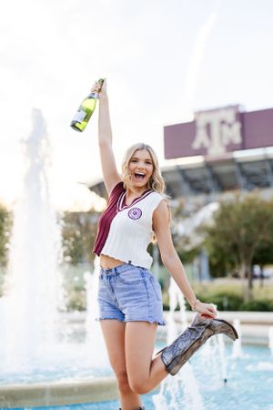 A college student in a Texas A&M crop top and denim shorts celebrates while holding a drink bottle near a fountain.