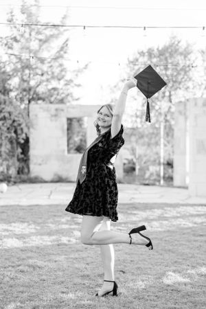 Black and white graduation photo showing someone in a floral dress holding up a graduation cap outdoors.