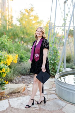 Graduate in black lace dress and burgundy top poses by garden with galvanized tub and yellow flowers in background.