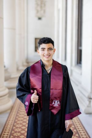 Graduate wearing black commencement gown with maroon Texas A&M stole gives thumbs up in elegant hallway.