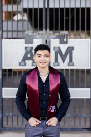 A student wearing a burgundy graduation stole and black shirt stands in front of Texas A&M University signage.