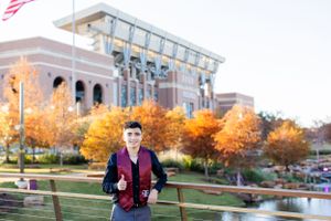 Student stands in front of campus building during autumn with colorful orange foliage and blue sky background.
