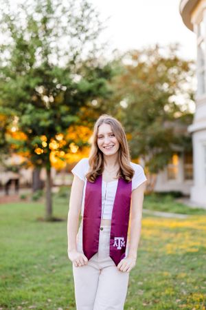 Graduate wearing a maroon and white graduation stole stands outdoors on campus lawn with trees and flowers in background.