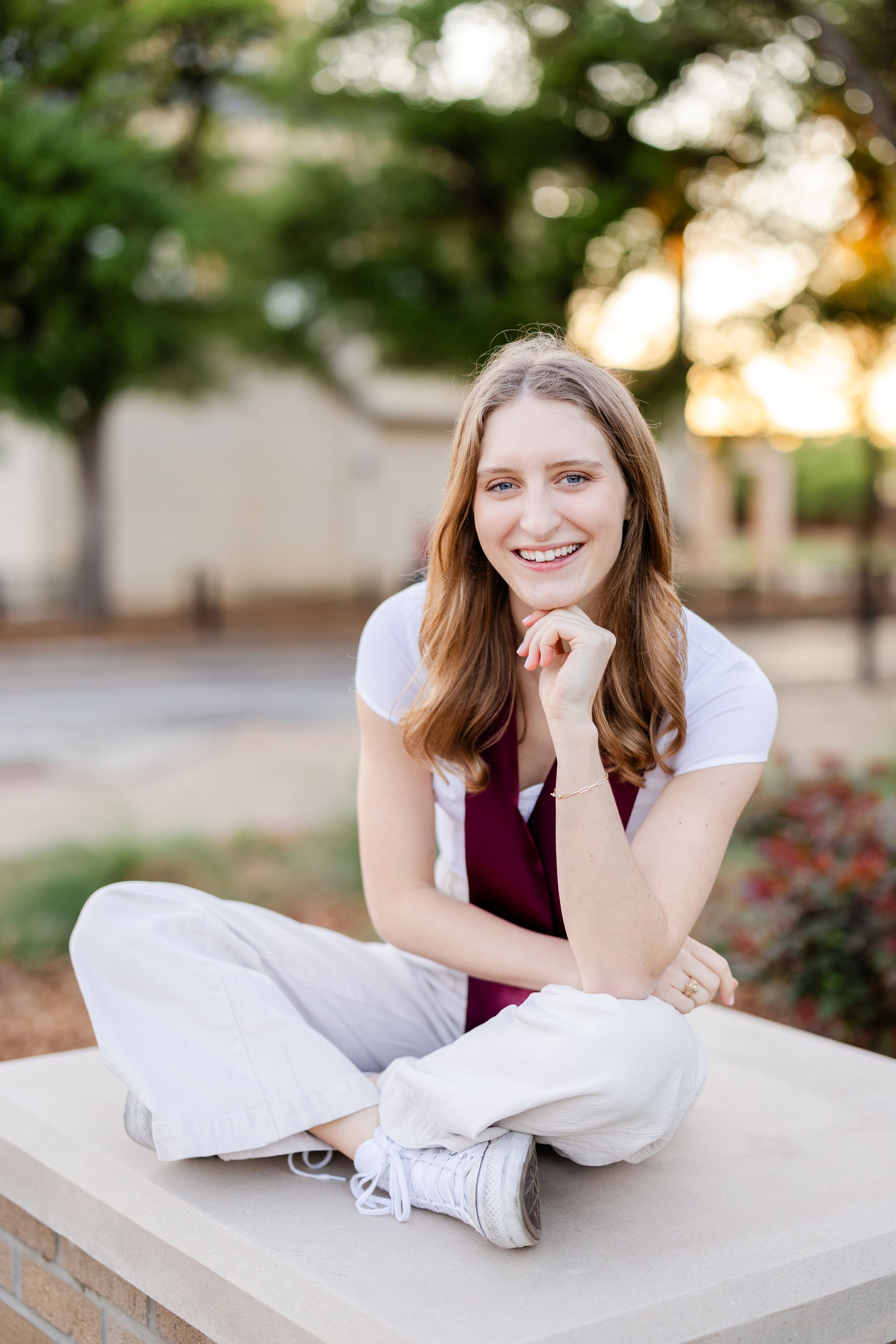 A person sits cross-legged in casual white and burgundy attire on a concrete ledge during golden hour.