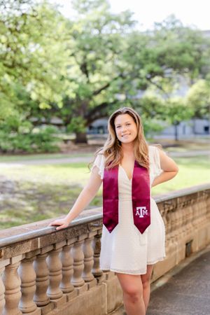 Graduate in white dress with maroon Texas A&M graduation stole poses by stone railing on sunny day.