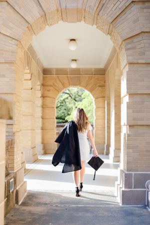 Graduate walks through stone archway corridor holding graduation gown and cap at university campus.