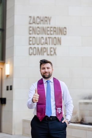 A graduate wearing maroon stole and blue tie gives thumbs up in front of the Zachry Engineering Education Complex.