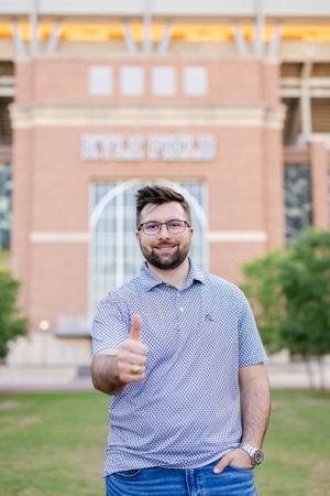 A person gives a thumbs up while standing outside a brick building wearing a blue polo shirt and jeans.