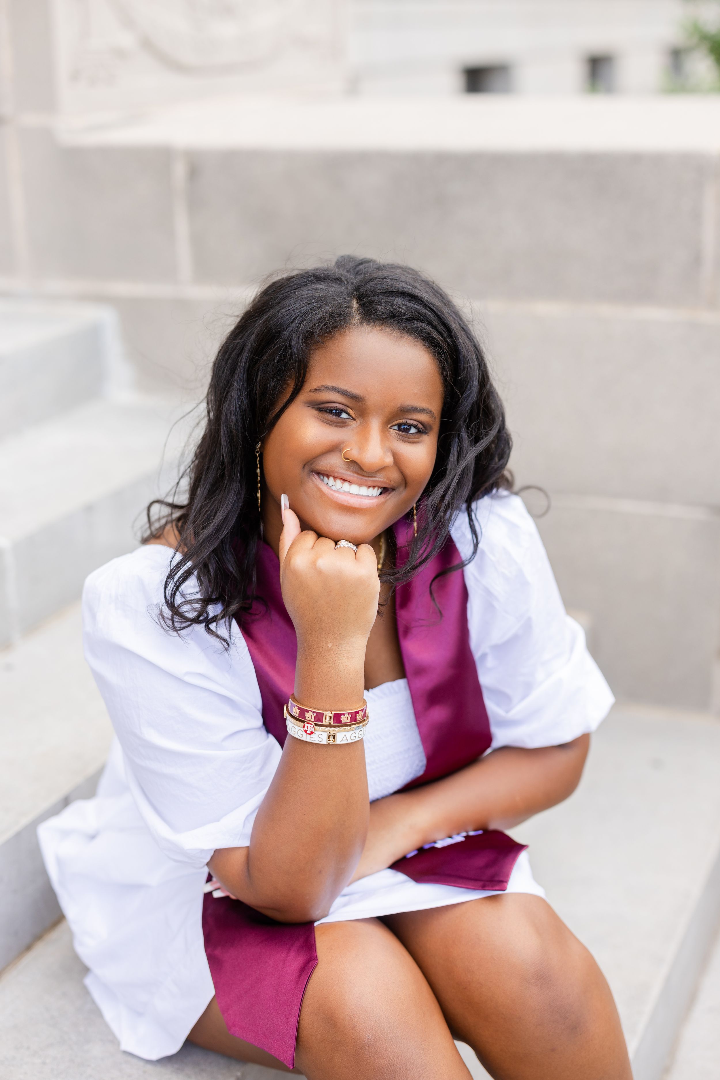 Student in a white blouse and burgundy dress sits on stone steps with a bright smile.