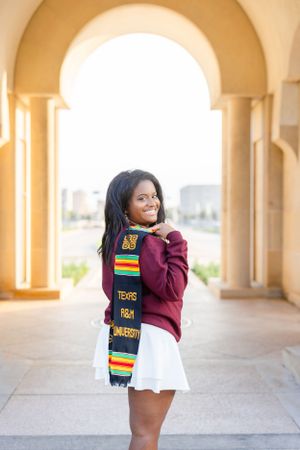 Graduate in burgundy sweater and white skirt poses under arches wearing traditional kente cloth graduation stole.