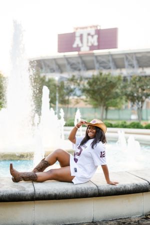 Student in western boots sits on fountain ledge with Texas A&M stadium in background near campus water feature.