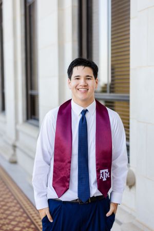A graduate wearing a maroon Texas A&M stole over white dress shirt and navy tie poses outdoors near columns.
