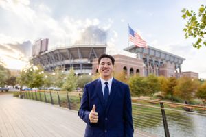 A person in a navy suit gives a thumbs up gesture outside a stadium with an American flag visible at sunset.