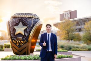 A large bronze police badge sculpture or monument stands in an outdoor plaza at sunset with a figure in a suit nearby.