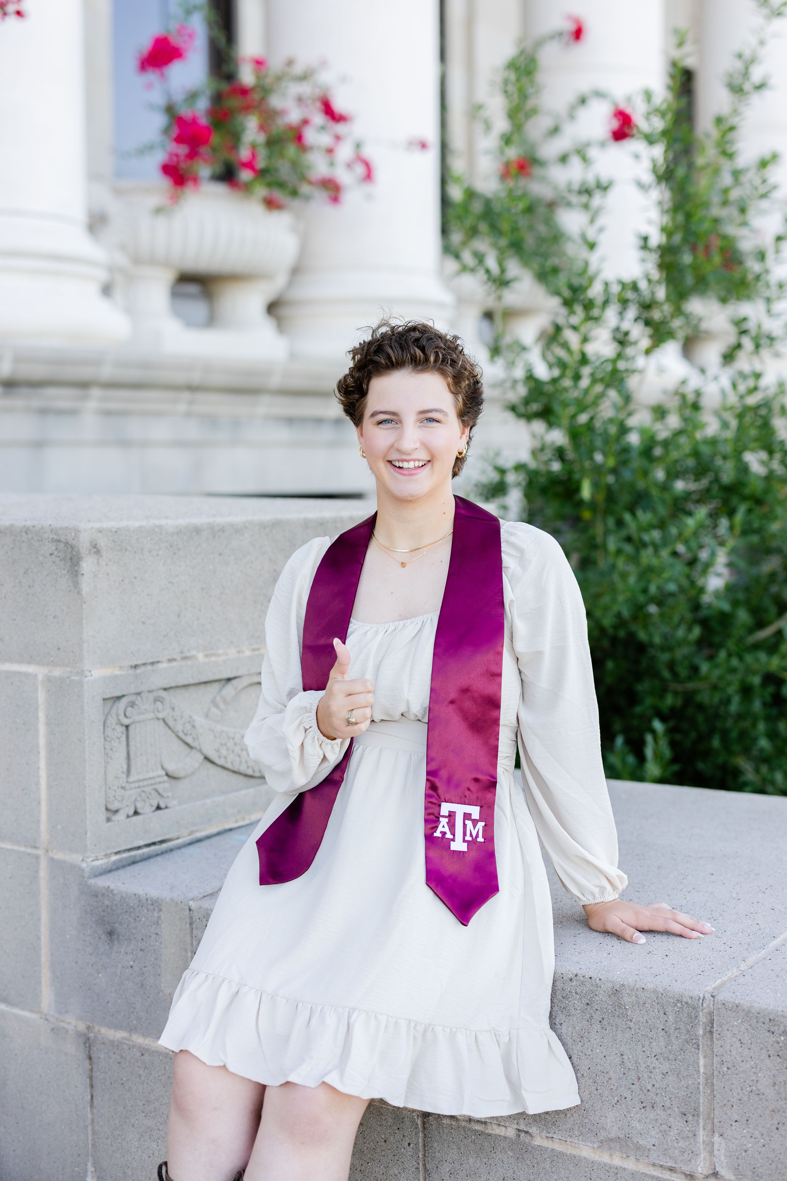 Graduate wearing a white dress and Texas A&M graduation stole poses outside near decorative stone columns and flowers.