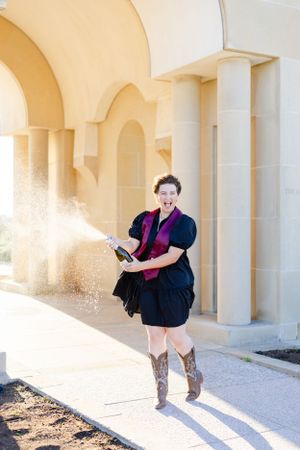 Figure in a dark dress and cowboy boots sprays champagne in front of an arched colonnade on a sunny day.