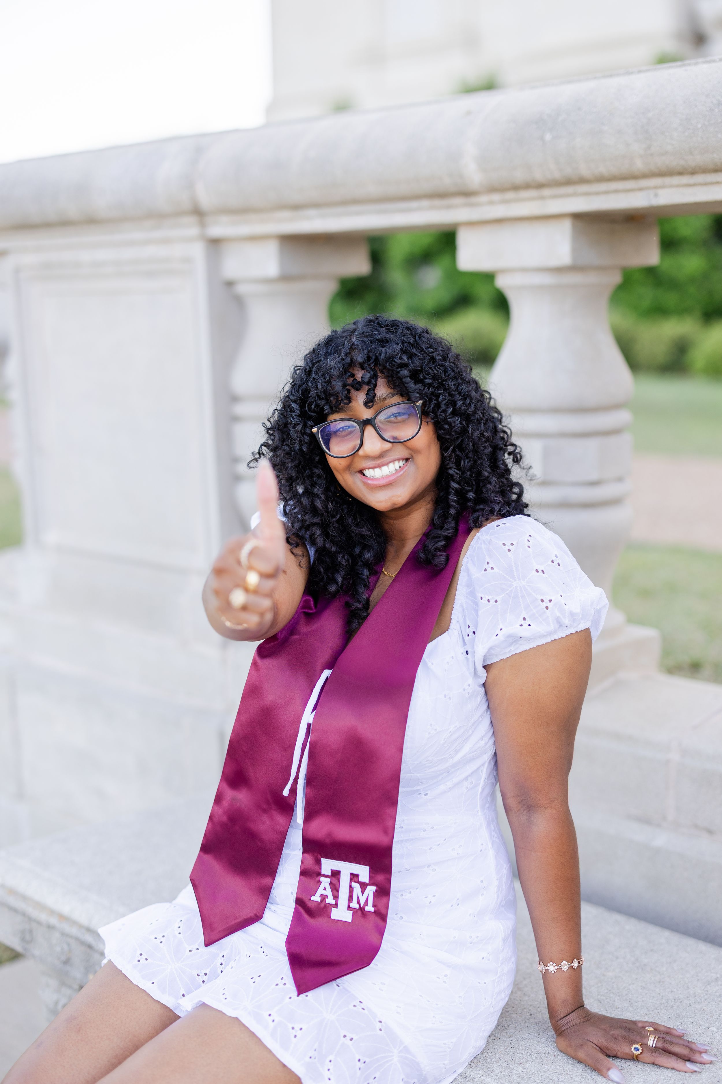 Graduate wearing burgundy Texas A&M graduation stole and white eyelet dress poses smiling on stone steps outdoors.
