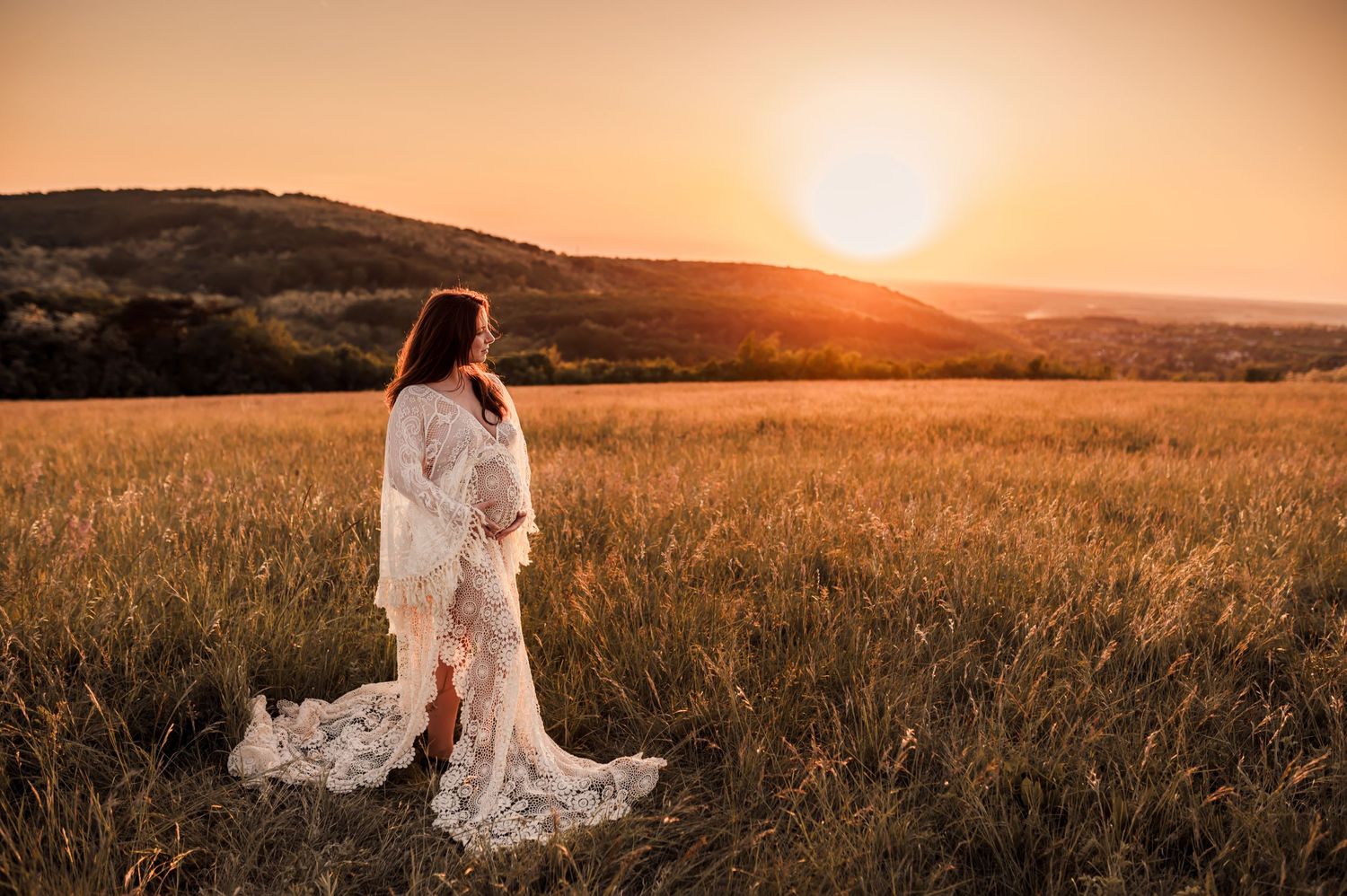 A figure in a flowing white lace dress stands in a golden wheat field at sunset with rolling hills in the background.