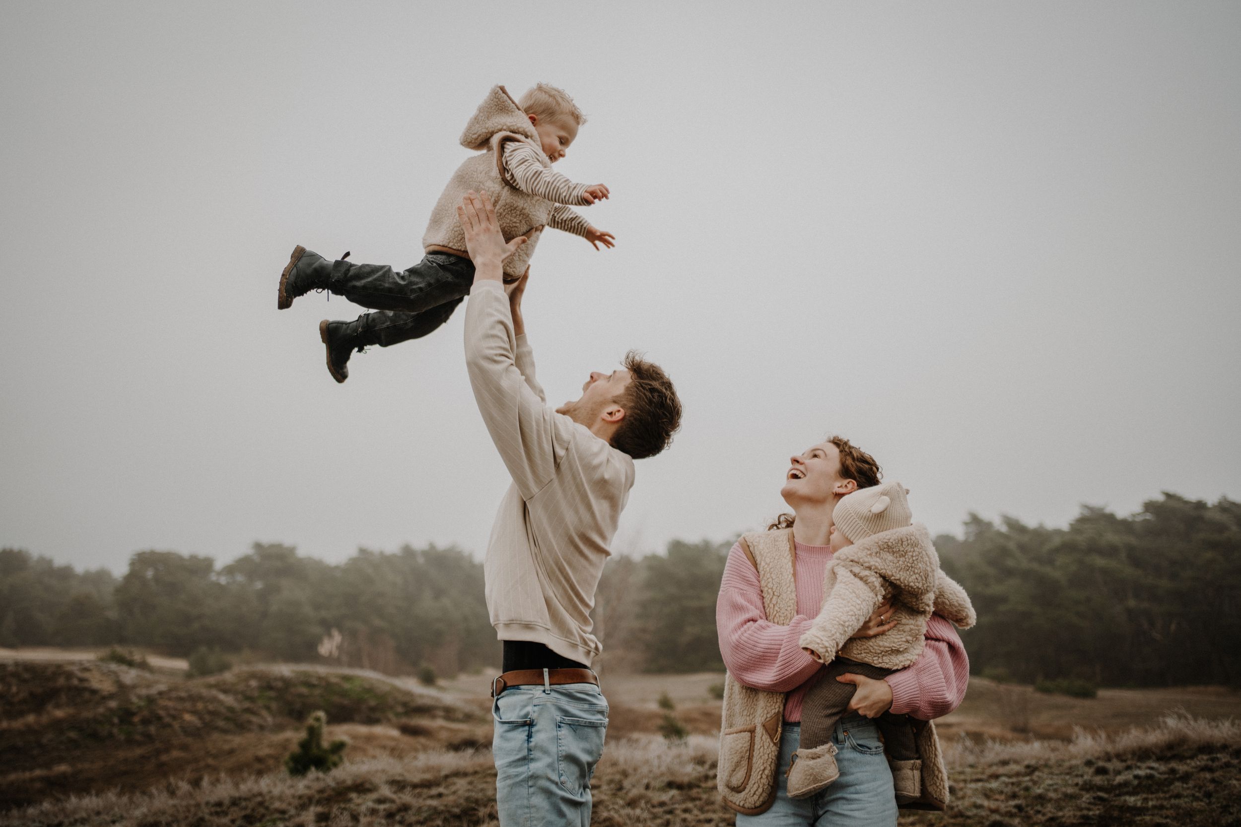 Playful family moment with child being lifted into the air against cloudy sky.