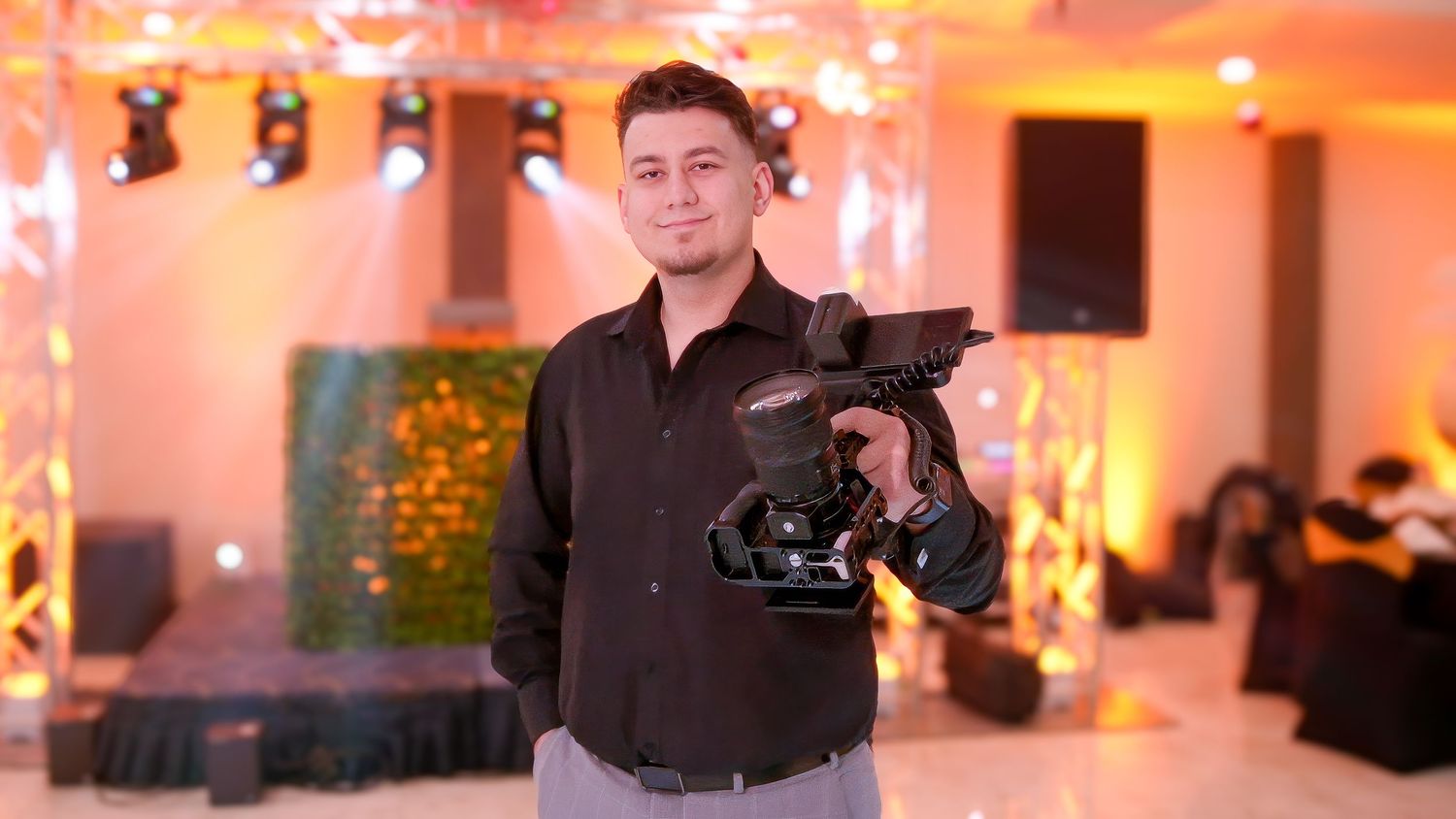 A professional videographer holds a camera rig at an indoor event with warm lighting.