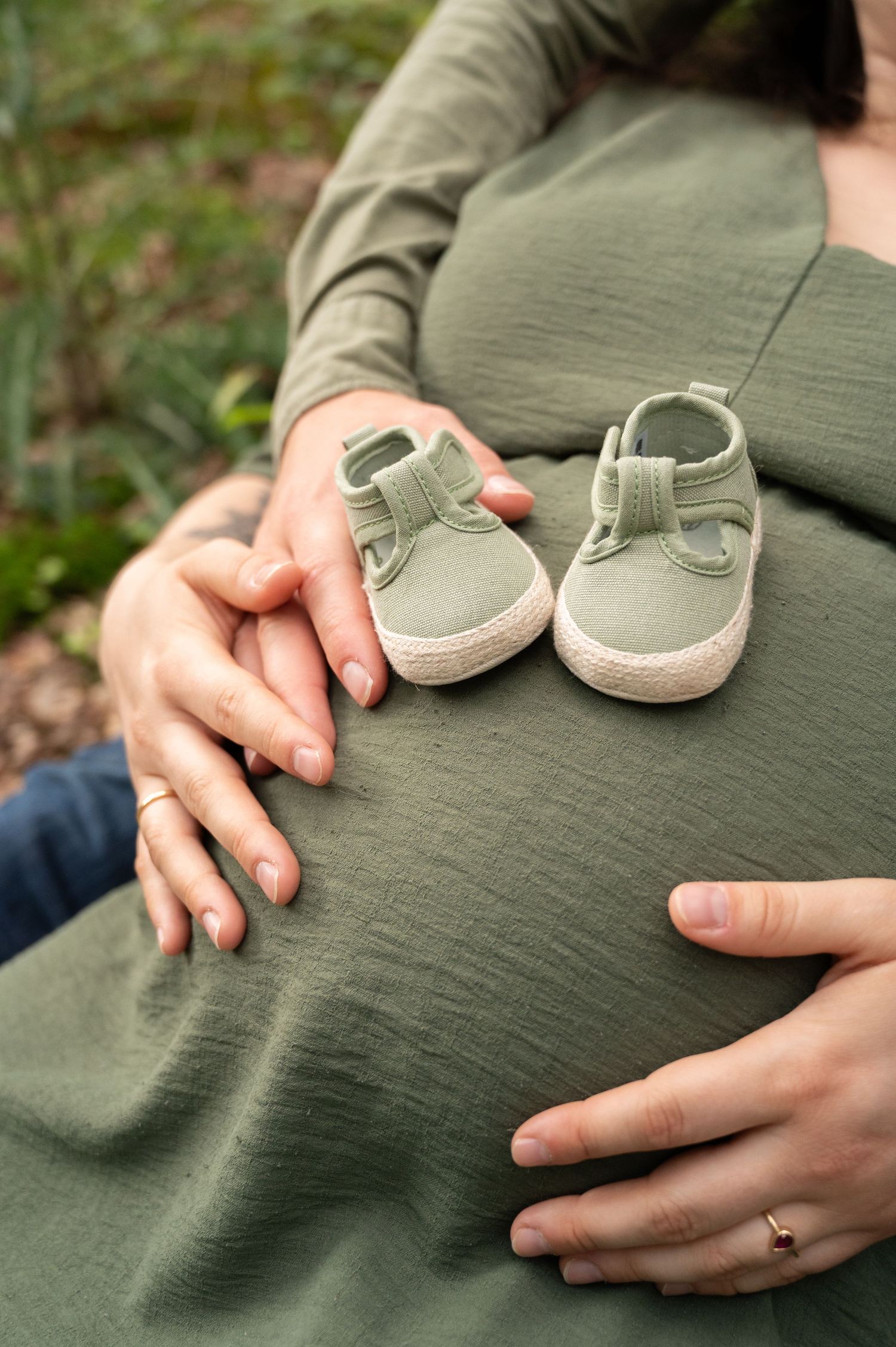 Détail du ventre d’une femme enceinte à Barvaux avec les mains des parents et des chaussons de bébé en extérieur naturel