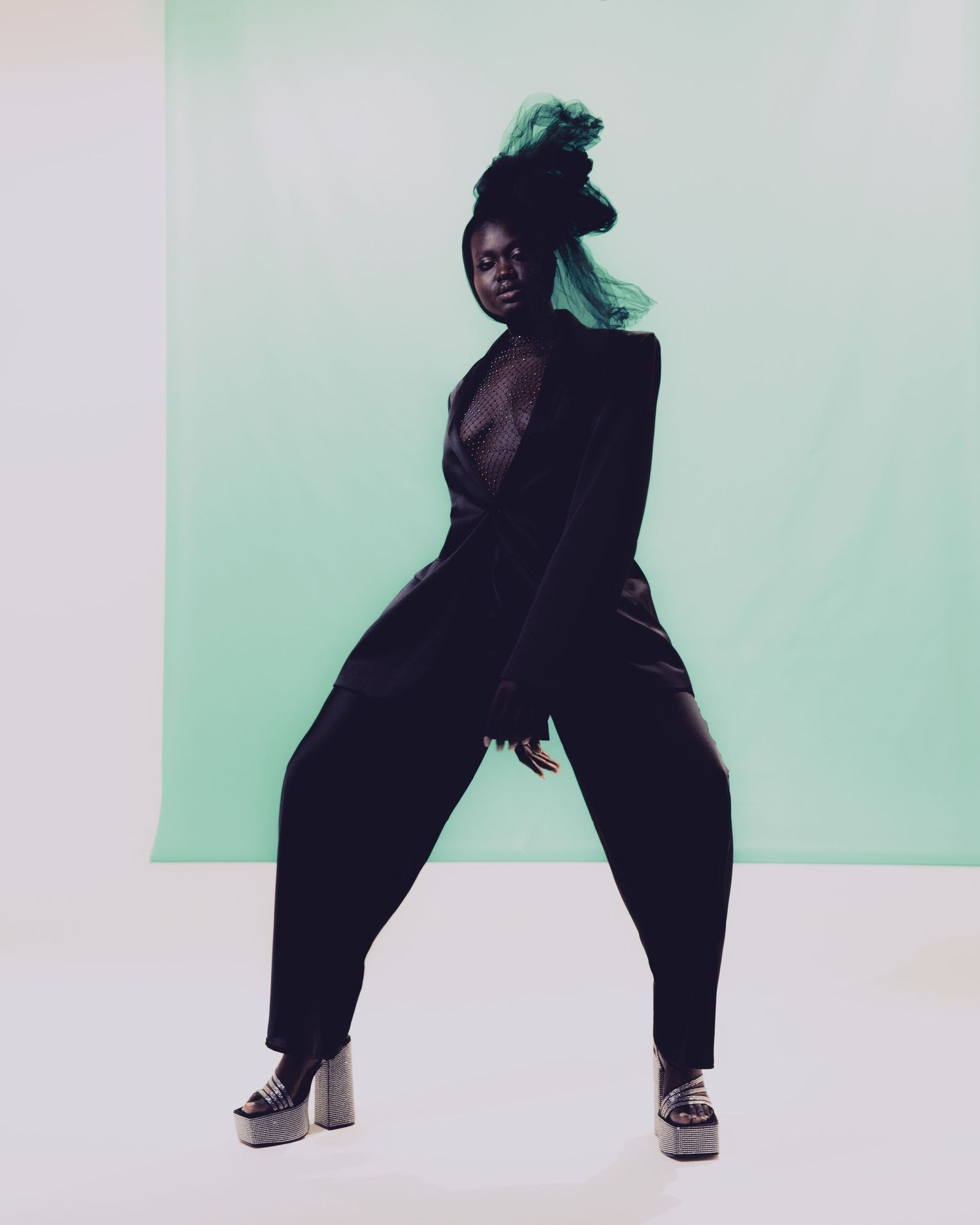 Dramatic fashion pose against mint green background wearing all-black ensemble.